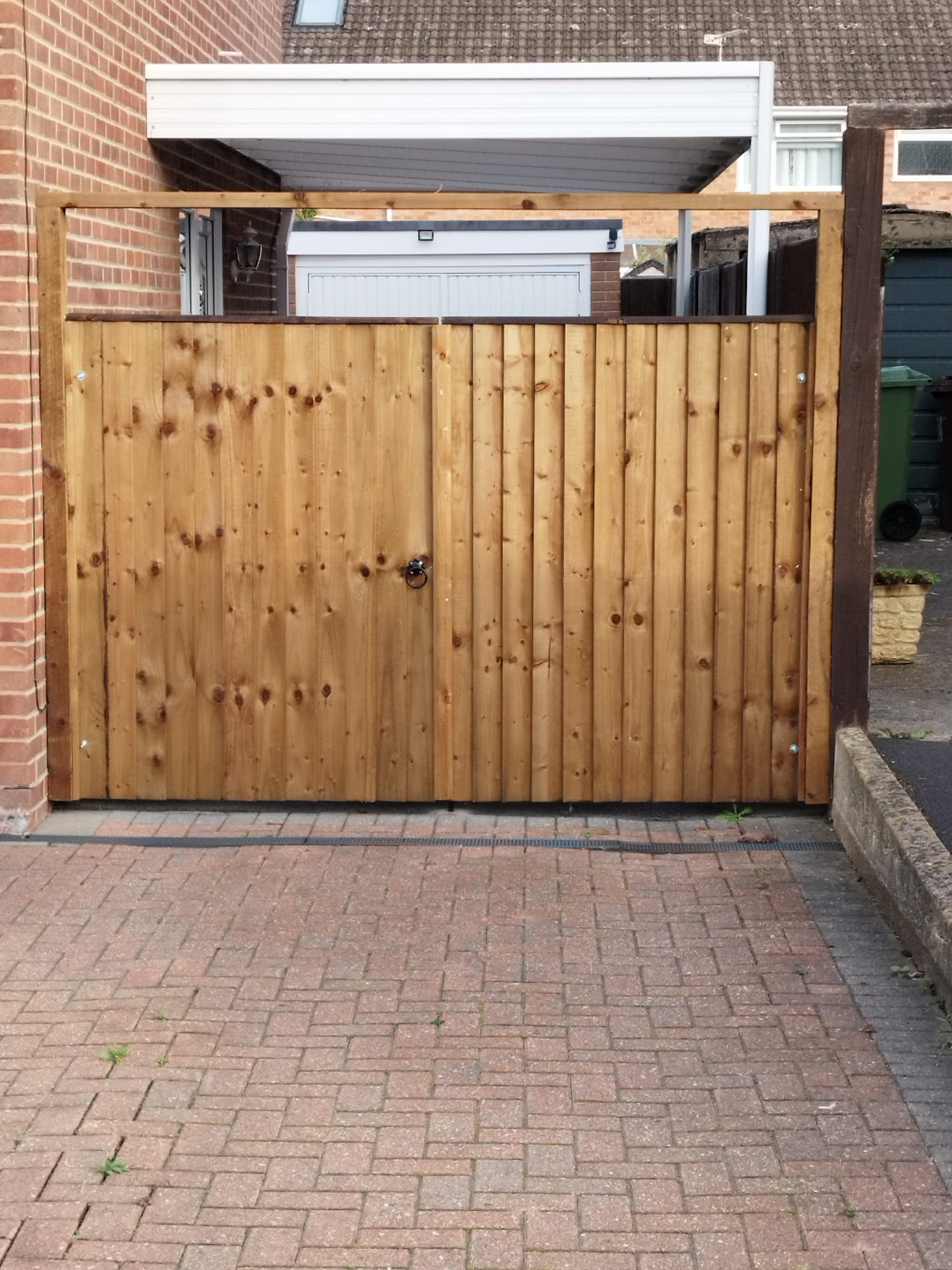 A wooden side gate made of vertical planks with a central handle, framed by wooden posts, set between a brick wall and a driveway, with a building and garage visible in the background.